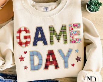 Könnte beinhalten: Cremefarbenes Sweatshirt mit den Worten "GAME DAY" in Patchwork-Buchstaben. Die Buchstaben weisen verschiedene Muster auf, darunter Karos, Punkte und Blumendrucke. Rote Herzen und Sterne akzentuieren das Design. Ein lässiges, sportliches Thema.