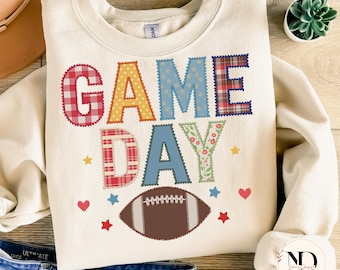 Könnte beinhalten: Cremefarbenes Sweatshirt mit den Worten "GAME DAY" in bunten Buchstaben im Patchwork-Stil. Eine braune Football-Grafik ist unter dem Text zentriert, mit kleinen Stern- und Herzakzenten. Das Sweatshirt hat einen Rundhalsausschnitt und lange Ärmel.