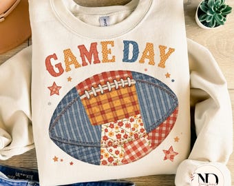 Könnte beinhalten: Cremefarbenes Sweatshirt mit den Worten "GAME DAY" in orangefarbenen und blauen Buchstaben. In der Mitte befindet sich eine Football-Grafik aus Patchwork-Stoff in Rot-, Blau- und Blumenmustern. Kleine Sternakzente umgeben den Football.