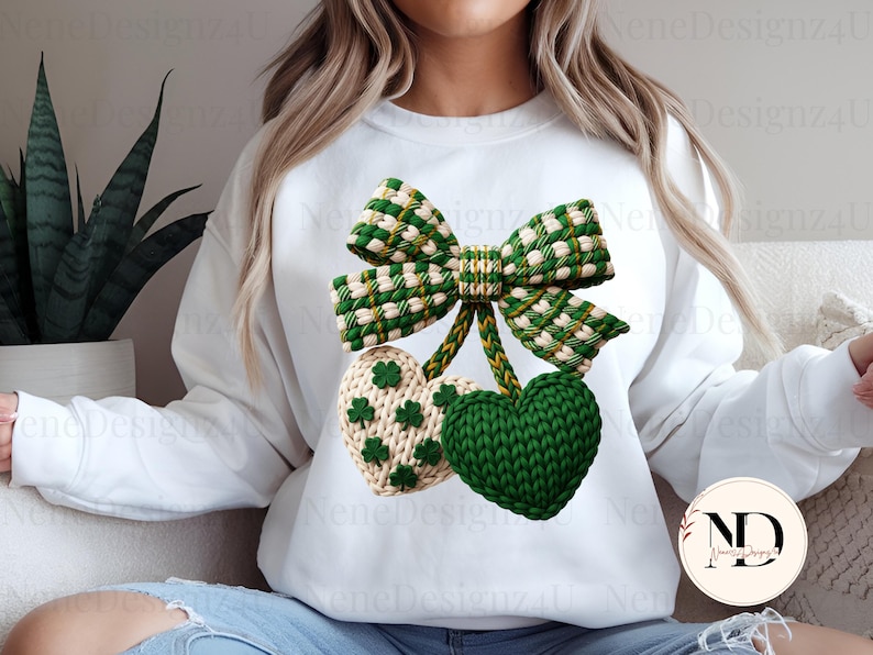 May include: A white sweatshirt featuring a green and white bow and two heart-shaped ornaments. One heart is green, the other is cream-colored with green shamrocks. The design is likely for St. Patrick's Day.