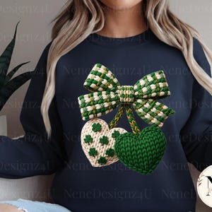 May include: Navy blue sweatshirt featuring a green and white bow and two heart-shaped ornaments. One heart is green, the other is white with green shamrocks. The design is a knitted style, perfect for St. Patrick's Day.