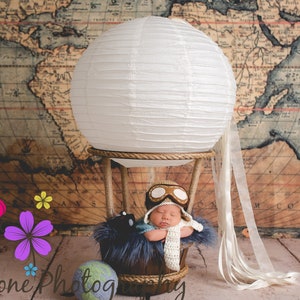 May include: A newborn baby wearing a pilot hat and goggles is sleeping in a white hot air balloon basket. The balloon is made of white paper and has a white ribbon tail. The baby is surrounded by a blue and white plush toy. The background is a vintage world map.
