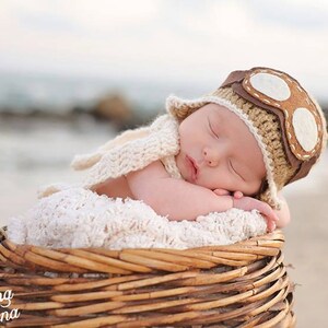 May include: A newborn baby wearing a brown and white crocheted aviator hat with goggles sleeps in a wicker basket. The baby is wrapped in a white crocheted blanket.