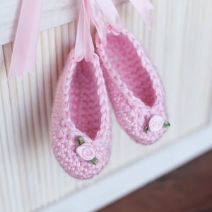 May include: A pair of pink crocheted baby shoes with white flower embellishments and green ribbon accents.