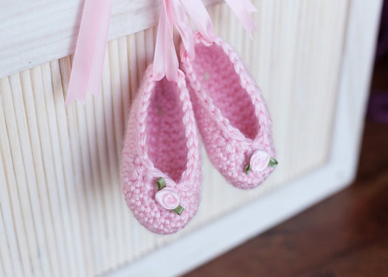 May include: A pair of pink crocheted baby shoes with white flower embellishments and green ribbon accents.
