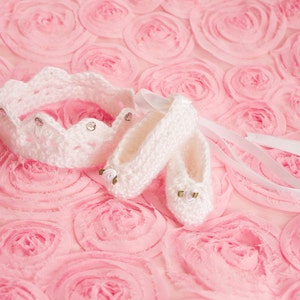 May include: White crocheted baby shoes and a crown headband with rhinestones on a pink background.
