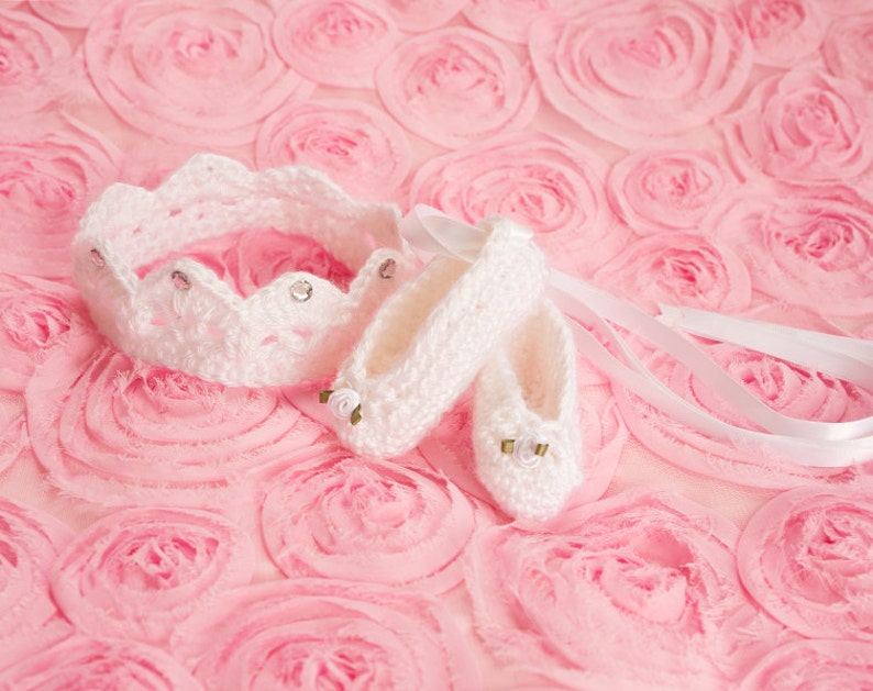 May include: White crocheted baby shoes and a crown headband with rhinestones on a pink background.