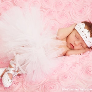 May include: A sleeping baby girl wearing a white tutu, white crocheted crown headband, and white ballet shoes. The baby is lying on a pink, rose-shaped blanket.