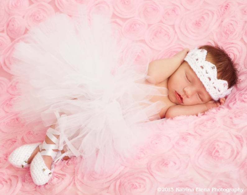 May include: A sleeping baby girl wearing a white tutu, white crocheted crown headband, and white ballet shoes. The baby is lying on a pink, rose-shaped blanket.