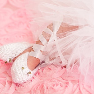 May include: A pair of white crocheted baby shoes with gold accents, worn by a baby wearing a white tulle skirt on a pink background.