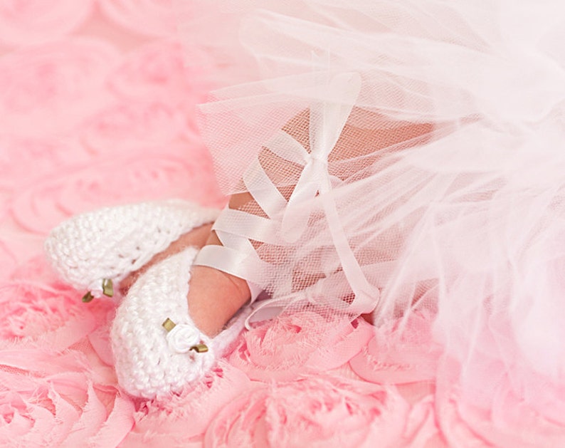 May include: A pair of white crocheted baby shoes with gold accents, worn by a baby wearing a white tulle skirt on a pink background.