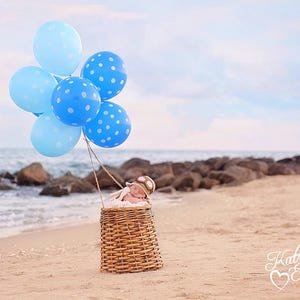 May include: A baby wearing a pilot hat is sleeping in a wicker basket. Blue and white polka dot balloons are tied to the basket and float in the air above the baby.