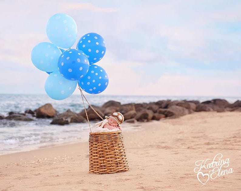 May include: A baby wearing a pilot hat is sleeping in a wicker basket. Blue and white polka dot balloons are tied to the basket and float in the air above the baby.