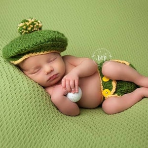 May include: A newborn baby wearing a green and yellow crocheted golf hat and matching diaper cover, holding a white golf ball. The baby is sleeping on a green knitted blanket.