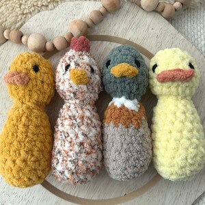May include: Four handmade crochet animal toys: a yellow chick, a chicken with a red comb, a gray and brown duck, and a yellow duck. Each has a beak and black eyes. They are arranged on a wooden tray with a beaded garland.