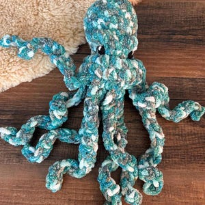 May include: A handmade plush octopus in shades of teal, white, and gray. The octopus has eight long, textured tentacles and black button eyes. The plush toy is made of soft, chunky yarn, perfect for a child's room or as a decorative item.