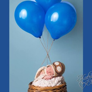 May include: A newborn baby wearing a brown aviator hat with goggles sleeps in a wicker basket, suspended by blue balloons.
