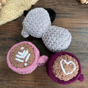 May include: Crocheted plush toys in the shape of a gray elephant with black ears and two coffee cups. One cup is pink with a white leaf design, and the other is burgundy with a white heart.