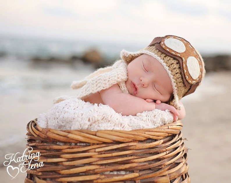 May include: A newborn baby wearing a brown and white crocheted aviator hat with goggles sleeps in a wicker basket. The baby is wrapped in a white crocheted blanket.