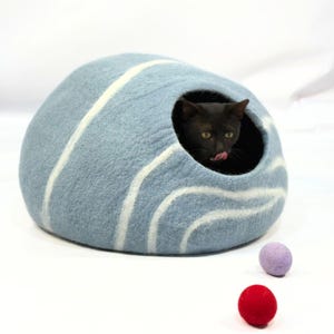 Può includere: Un letto per gatti in feltro blu e bianco a forma di cupola con un gatto nero che sporge dall'ingresso. Ci sono due piccole palline di feltro rosso e viola sulla superficie bianca.
