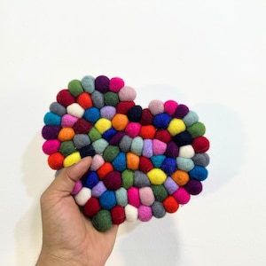 Può includere: Un sottopentola a forma di cuore fatto a mano, composto da numerose palline di feltro multicolore. I colori includono rosso, rosa, blu, giallo e verde. Il sottopentola è tenuto in mano su uno sfondo bianco.