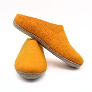May include: A pair of mustard yellow felted wool slippers with a slip-on design. The slippers feature a rounded toe, a low-cut opening, and a light grey sole that is stitched to the upper portion of the footwear with visible thread detailing.