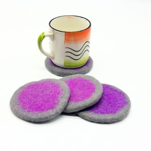 May include: A white mug with orange and green accents sits on a grey coaster. Three additional round coasters with a purple centre and grey border are in the foreground. The coasters are made of felt.