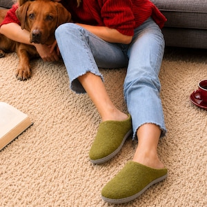 Puede incluir: Un par de zapatillas de lana afieltrada en color verde oliva con punta redondeada y suela plana gris. Las zapatillas presentan un diseño sencillo de tipo zueco con talón cerrado y una superficie exterior suave y texturizada.
