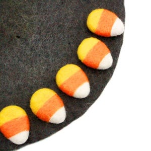 Può includere: Un cerchio di feltro grigio scuro decorato con sei forme di caramelle di mais. Ogni caramella di mais è bianca, arancione e gialla. Il feltro ha un aspetto leggermente strutturato e le caramelle di mais sono disposte in un motivo curvo.