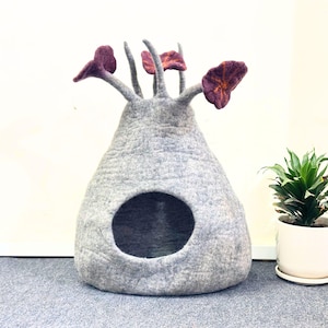 Pode incluir: Uma cama para animais de estimação de feltro cinza em forma de cápsula com uma entrada redonda. O topo tem caules com detalhes roxos em forma de flor. Uma planta em vaso está à direita.