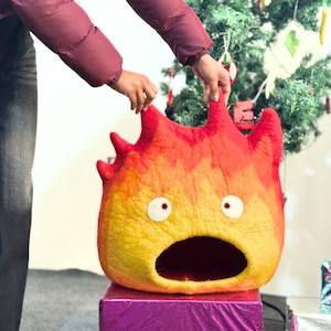 Peut inclure: Un lit pour animaux de compagnie en feutre en forme de démon de feu de dessin animé, avec des couleurs rouge, orange et jaune. Le lit a une grande bouche ouverte et des détails d'yeux blancs. Il est placé sur une boîte violette.