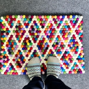 Puede incluir: Una alfombra rectangular multicolor de bolas de fieltro con un patrón de rejilla blanca. La alfombra presenta una vibrante gama de colores, incluyendo rojo, amarillo, verde, azul y morado. La alfombra mide aproximadamente 60 cm de ancho y 90 cm de largo. Se ven pies con calcetines estampados.
