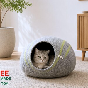 May include: A grey felted wool cat cave with a rounded dome shape and a large circular opening. The exterior features two thin, diagonal lime green stripes and a small white brand tag. A grey tabby cat is resting inside the enclosed structure.