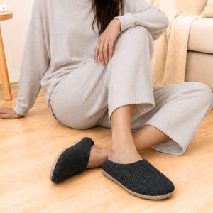 Puede incluir: Un par de zapatillas de lana afieltrada en color gris carbón oscuro con punta redondeada y suela gris claro. Las zapatillas presentan un diseño sin cordones con la parte trasera abierta, ofreciendo un estilo sencillo y funcional.