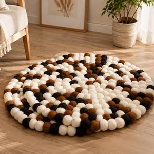 May include: A circular rug constructed from numerous small, densely packed felt balls in a repeating pattern of cream, brown, and black colours. The individual spheres are arranged in concentric circles to form a textured, thick, round floor covering.