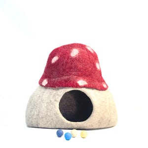 May include: A mushroom-shaped pet house made of felt, featuring a red cap with white spots and a light gray base with a circular entrance. Several small felt balls in blue, yellow, and white are at the base.