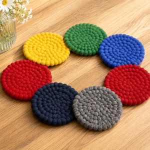 May include: A set of seven circular coasters made from small felted wool balls in various colors, including yellow, green, blue, red, dark blue, and gray, arranged in a circular pattern on a light brown wooden surface near a glass vase.