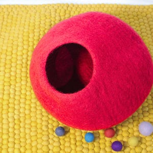 May include: A bright red felt cat cave with a large circular opening, set on a textured yellow mat. Several small, colorful felt balls in blue, purple, red, and yellow are scattered around the cave. This cozy cat bed is a vibrant addition to any home.