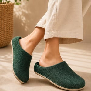 Puede incluir: Un par de zapatillas de lana afieltrada de color verde oscuro con punta redondeada, diseño de talón abierto y suela texturizada de color claro. Las zapatillas presentan costuras visibles a lo largo del borde donde la parte superior se une a la suela.