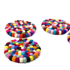 May include: A collection of colorful felt ball coasters. Each coaster is round and made of tightly packed, small felt balls in various colors, including pink, blue, yellow, and green. The coasters are designed to protect surfaces from heat and moisture.
