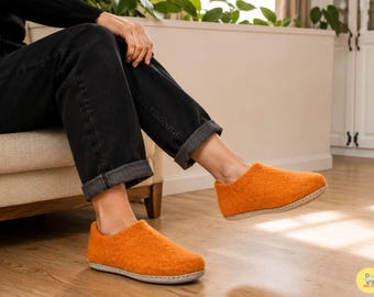 May include: A pair of bright orange felted wool slippers featuring a rounded toe, a low-cut ankle opening, and a light-colored stitched sole. The slippers have a textured, soft appearance and are worn with dark charcoal gray cuffed denim jeans.