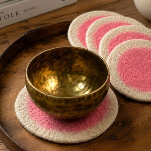 Può includere: Una ciotola tibetana in ottone metallico con una texture martellata poggia su un sottobicchiere rotondo in feltro color bianco sporco con un centro circolare rosa vivace. Cinque sottobicchieri identici sono impilati su un vassoio in legno scuro.