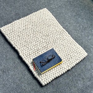 May include: A rectangular, off-white felt rug with a dense, textured surface. A closed, navy blue book with glasses on top rests on the rug. The rug is on a gray surface.