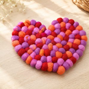 May include: A heart-shaped trivet constructed from numerous small, round felt balls in vibrant shades of red, orange, purple, and pink. The individual felt spheres are densely packed together to form a solid, textured heart shape with a soft appearance.