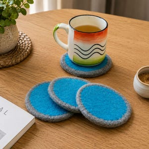 May include: A set of four round felt coasters, each featuring a vibrant blue center surrounded by a thick grey border. One coaster supports a ceramic mug with a white, orange, and green gradient design and black wavy lines, while three others lie flat.