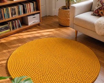 May include: A circular rug constructed from numerous small, mustard yellow felted wool balls arranged in a dense, uniform pattern. The rug lies flat on a wooden floor, showcasing a consistent spherical texture across its entire surface area.
