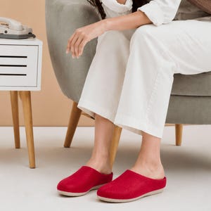 Puede incluir: Un par de zapatillas de fieltro rojo sin talón con punta redondeada y suela de color claro. Las zapatillas presentan un material superior rojo sólido y un diseño sencillo y abierto en la parte posterior para facilitar su uso. Suela plana.