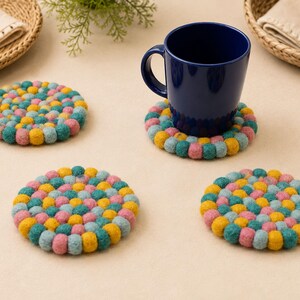 May include: Three circular coasters made of small, multicolored felt balls in shades of teal, yellow, and pink. One coaster supports a solid dark blue ceramic mug. The coasters are arranged on a flat surface, showcasing a dense, textured pattern.