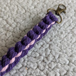 Puede incluir: Una correa trenzada para llavero en tonos morado y rosa, con un cierre de color bronce. La correa est&aacute; hecha de un cord&oacute;n grueso y tejido, creando una apariencia texturizada. El fondo es una superficie suave y blanca.