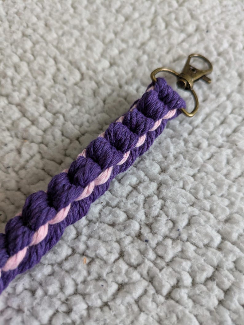 Puede incluir: Una correa trenzada para llavero en tonos morado y rosa, con un cierre de color bronce. La correa est&aacute; hecha de un cord&oacute;n grueso y tejido, creando una apariencia texturizada. El fondo es una superficie suave y blanca.